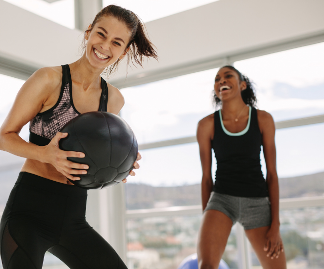 Two women laughing and exercising with a medicine ball in a bright fitness studio with large windows showing a scenic view.