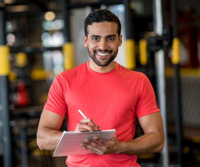 A smiling personal trainer in a red t-shirt holding a clipboard in a gym with exercise equipment in the background. (PT Membership Promotions)