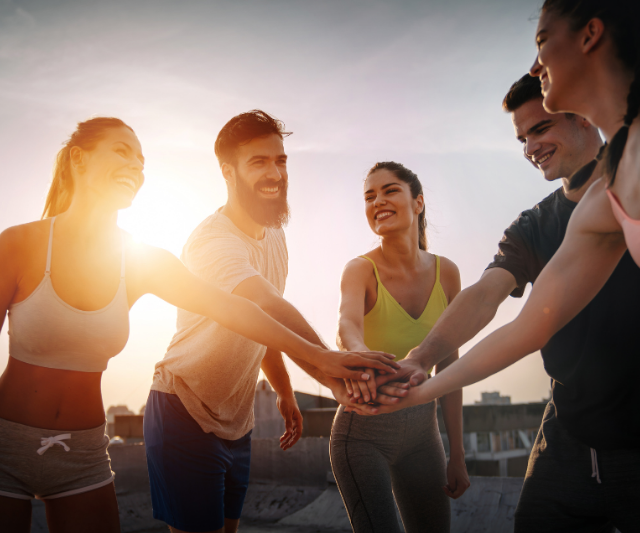 Eine Gruppe lächelnder Menschen in Trainingskleidung, die ihre Hände zu einem Stapel zusammenlegen, als Geste der Teamarbeit und Motivation, im Freien bei Sonnenuntergang.