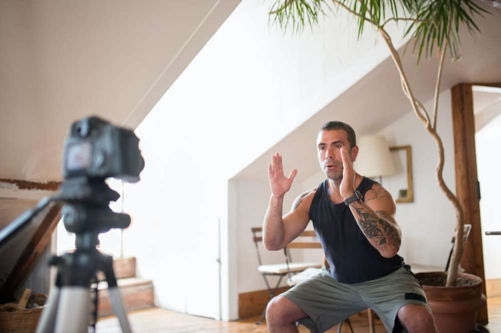 A man in athletic wear recording a fitness video at home using a camera on a tripod, demonstrating an exercise with his hands raised in front of him.