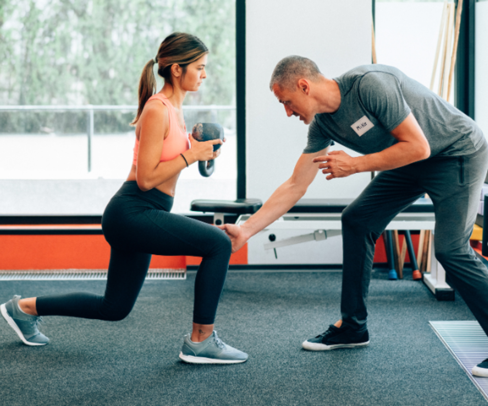 Ein Trainer unterstützt eine konzentrierte Frau, die in einem Fitnessstudio Ausfallschritte mit Kurzhanteln macht.