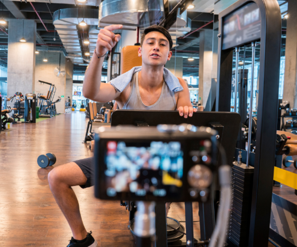 Male fitness trainer pointing at camera, with gym equipment in the background.