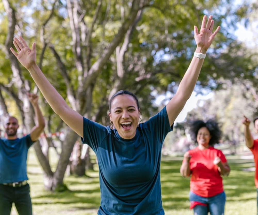 A group of people joyfully exercising outdoors, with a woman in the foreground enthusiastically raising her hands.