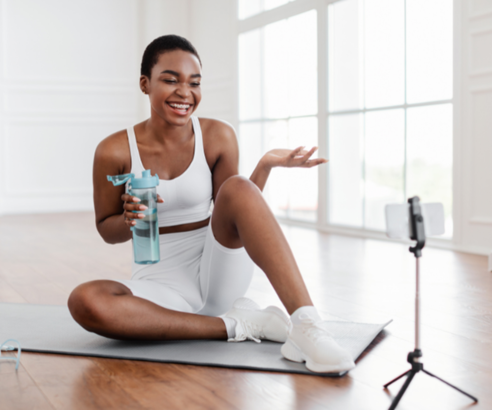 A woman in fitness attire sitting cross-legged on a yoga mat with a water bottle, smiling and interacting with a smartphone on a tripod, suggesting a fitness influencer or a virtual workout session.