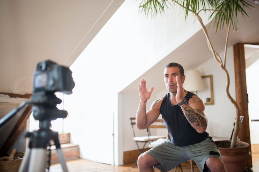 A man in casual attire demonstrating a martial arts stance while recording a fitness training video in a cozy, plant-filled indoor setting.