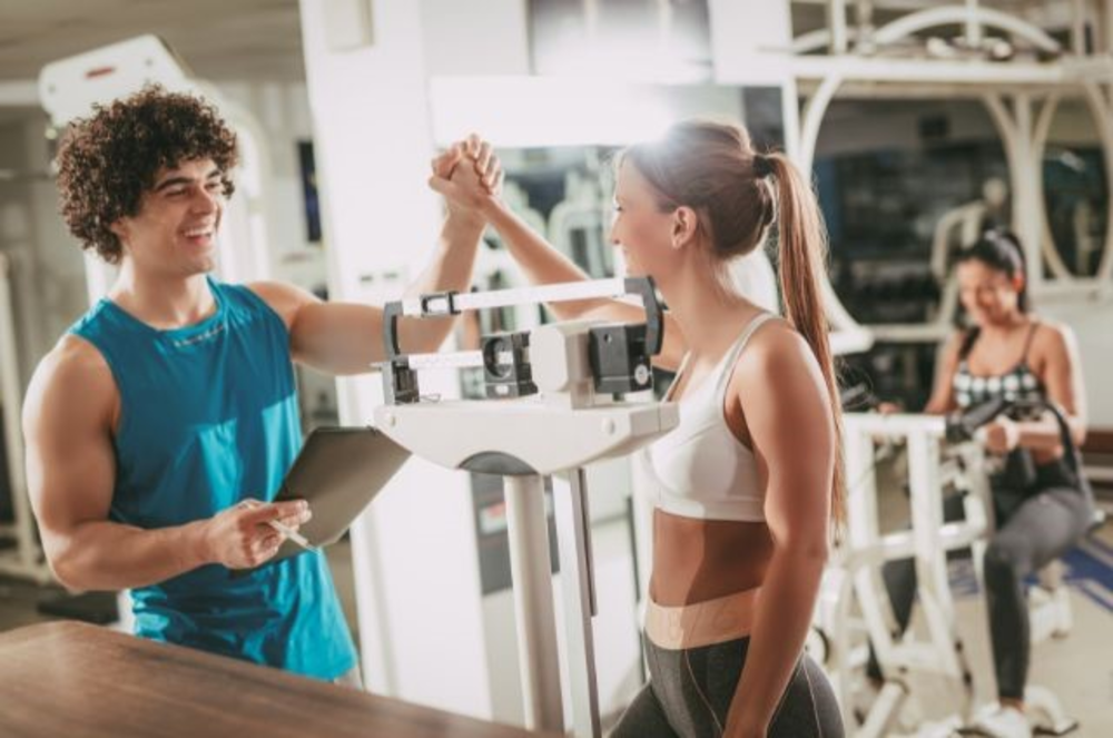 Un entrenador personal y una clienta celebrando el progreso con un choque de manos junto a la báscula en el gimnasio.