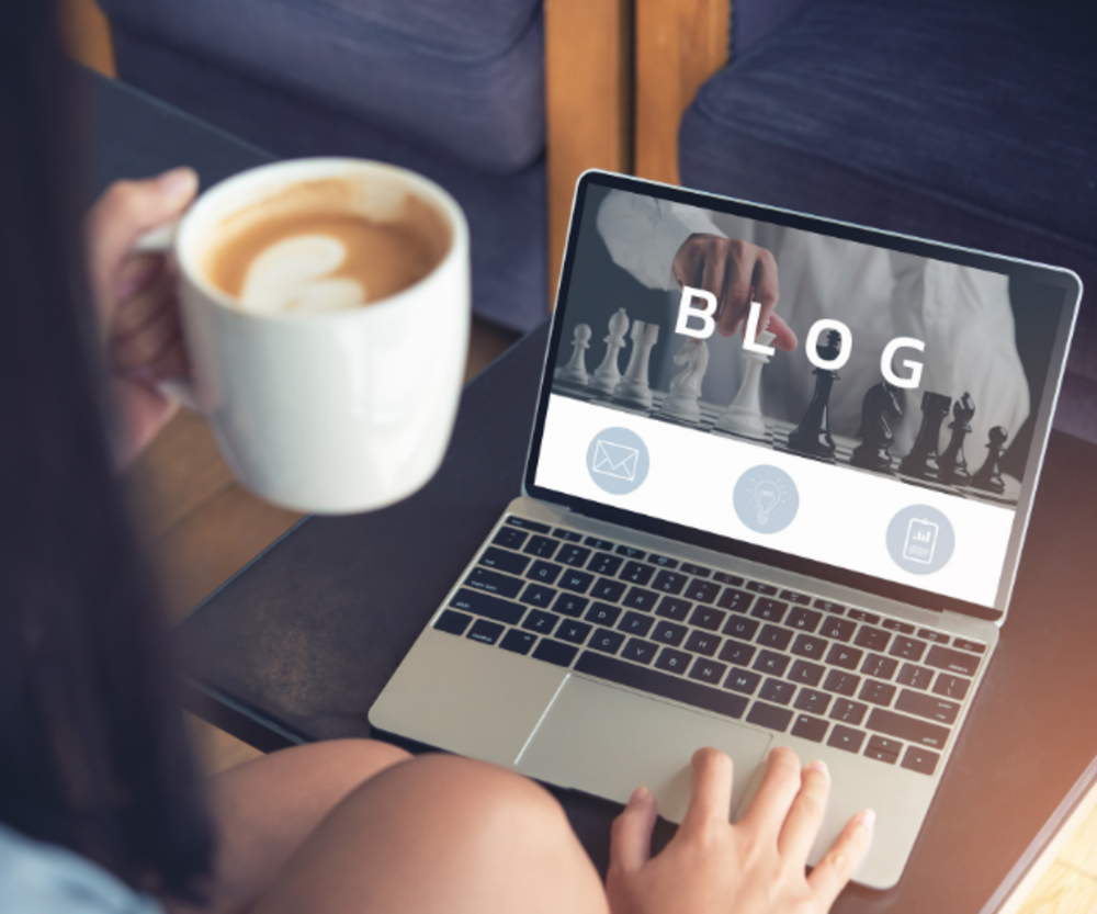 A lifestyle image of a person holding a coffee mug and using a laptop with a &quot;BLOG&quot; screen, depicting a relaxed blogging or content creation session.