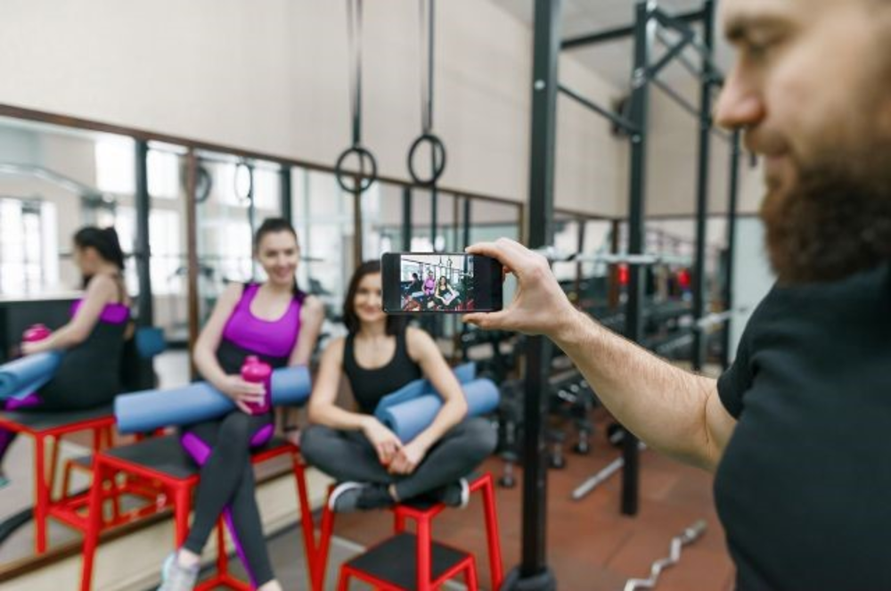 Una persona tomando una foto de dos clientas sentadas y sonriendo en un gimnasio, capturando un momento social después del entrenamiento.