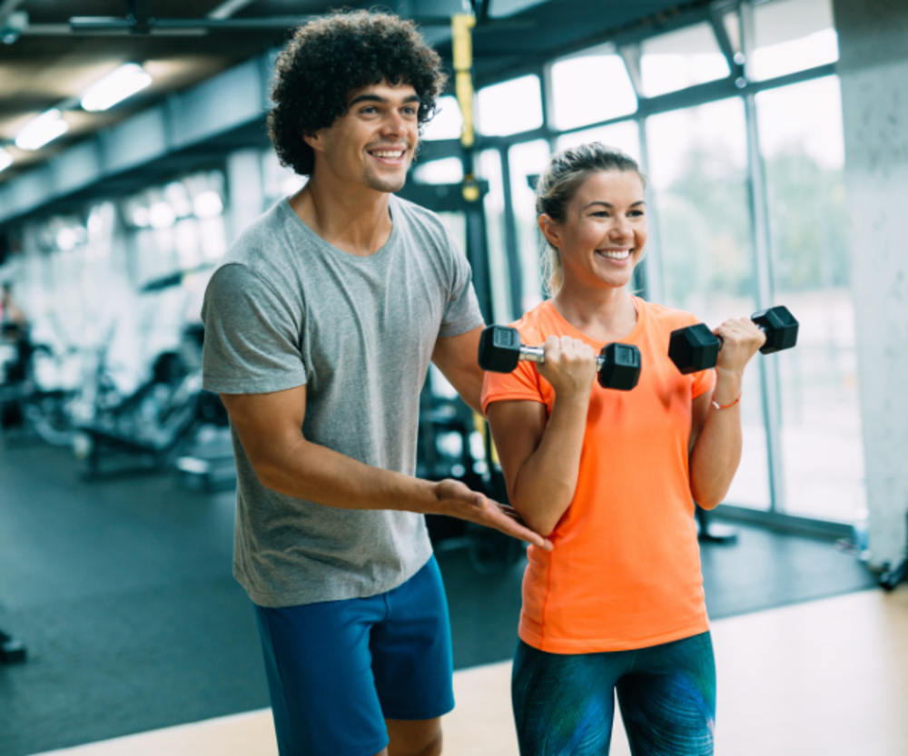 Un entrenador personal asistiendo a una mujer en ejercicios con mancuernas en un gimnasio, ambos sonriendo y enfocados en el entrenamiento.