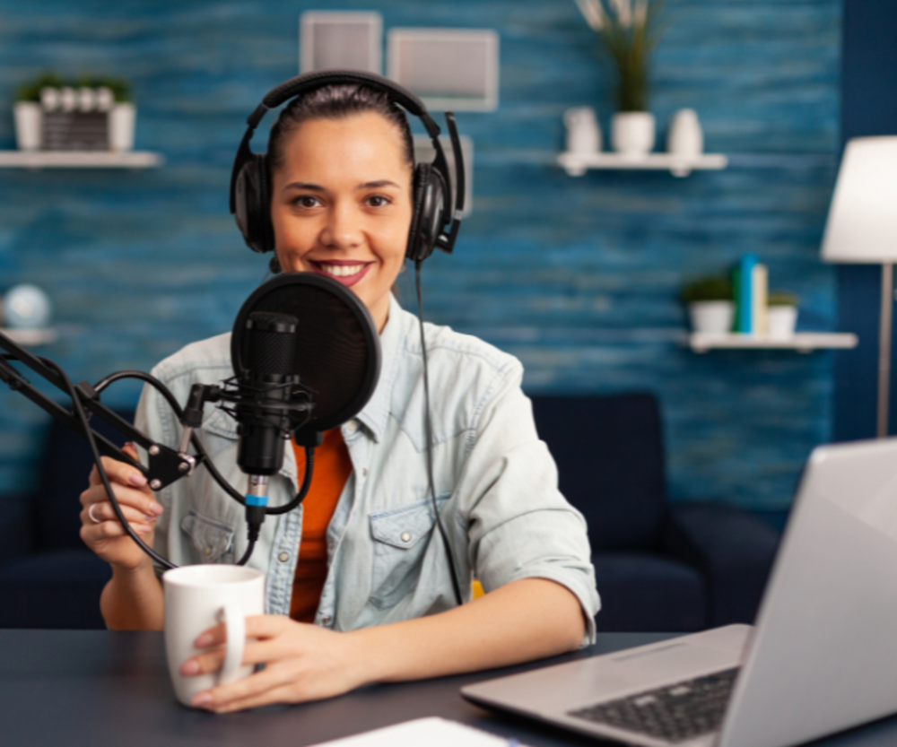 Eine lächelnde Frau trägt Kopfhörer und spricht in ein Mikrofon, vermutlich moderiert sie einen Podcast oder eine Webshow.