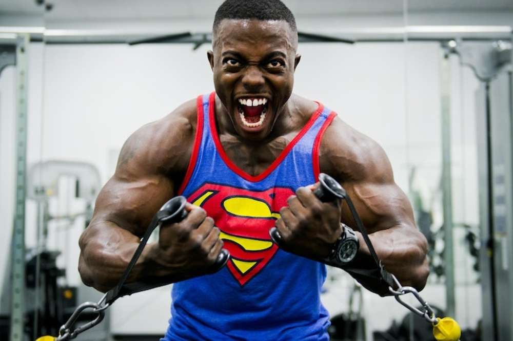 Un hombre musculoso con una camiseta sin mangas de Superman realizando un ejercicio con cables, mostrando una energía intensa y fuerza.