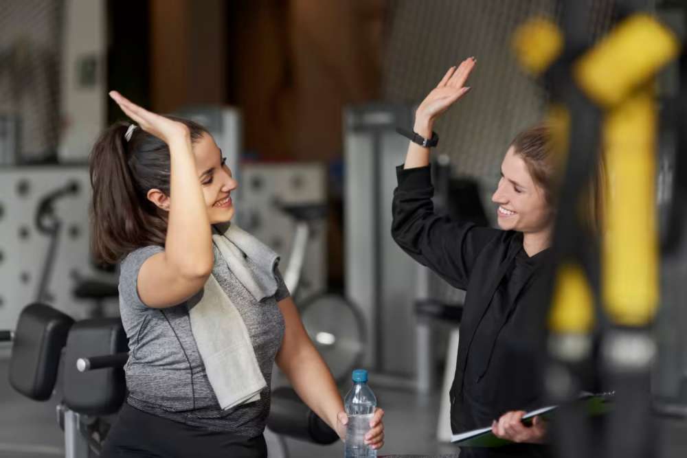 Two women in a gym smiling and giving a high five, with workout equipment in the background.