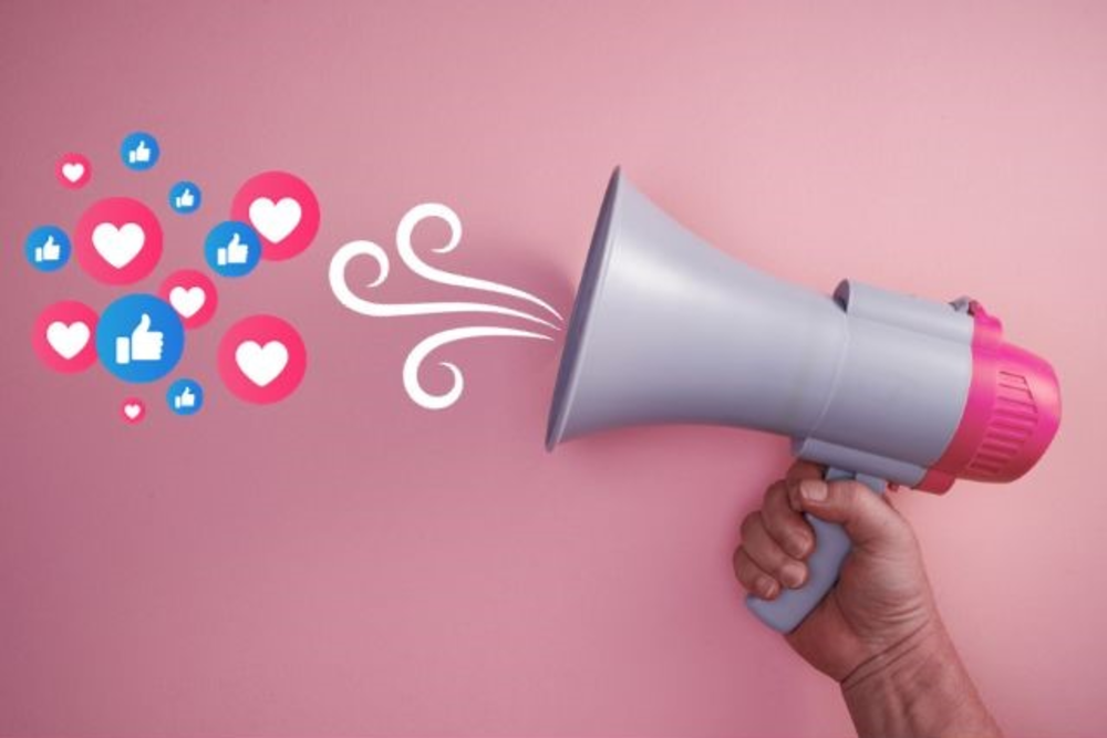 A hand holding a megaphone with icons of likes and hearts coming out of it, against a pink background, symbolizing social media engagement.