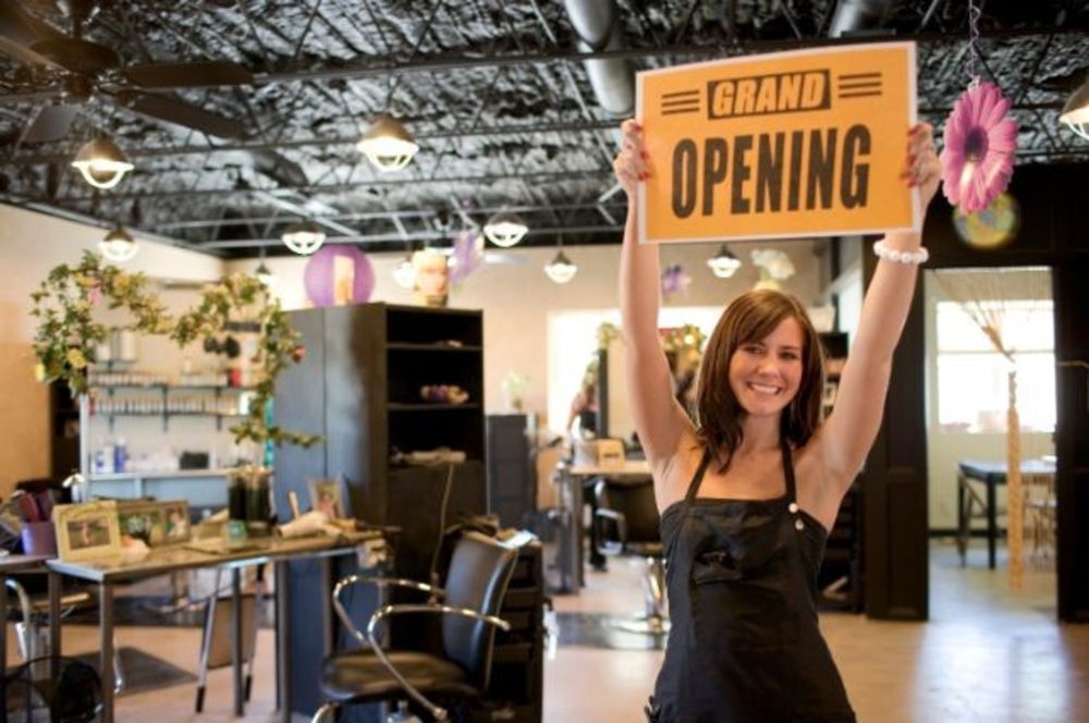 A woman happily holding a "Grand Opening" sign inside a new, stylish salon with empty chairs and decorative elements.