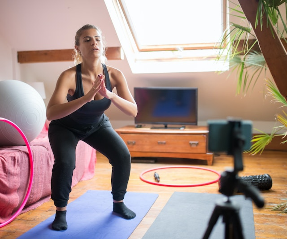 A woman in a yoga pose on a mat in a home setting with fitness equipment around, filming herself with a smartphone on a tripod, likely for an online training session or fitness vlog.