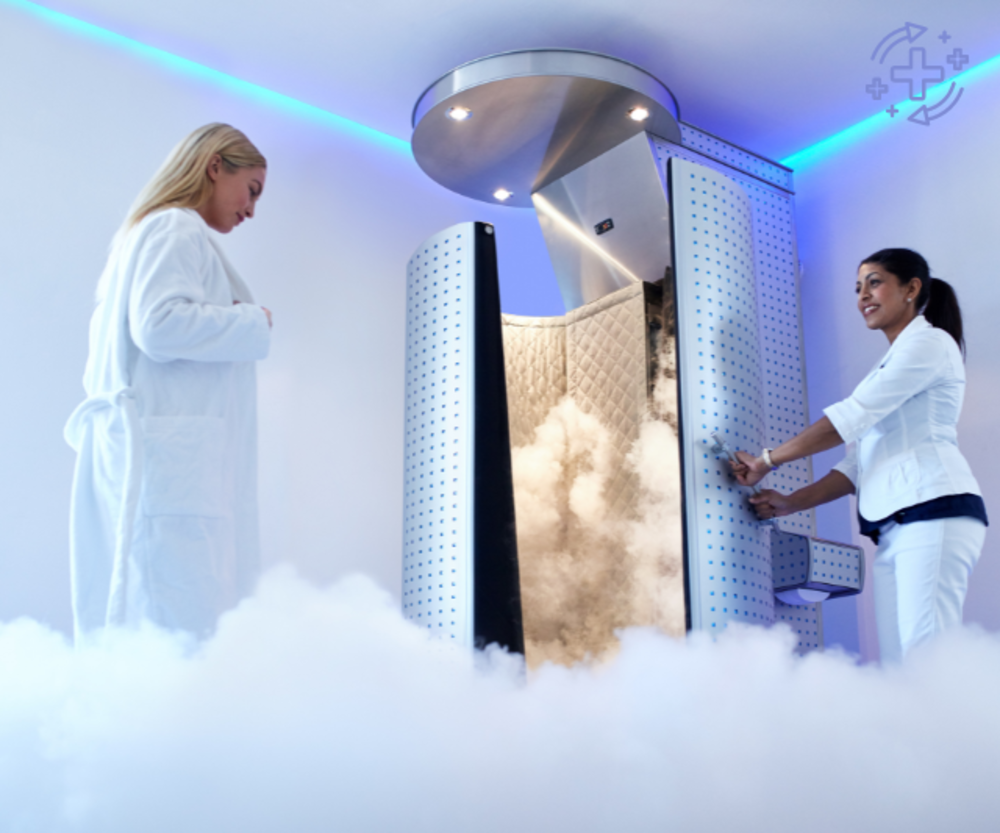 A woman in a white robe standing near a cryotherapy chamber emitting mist, assisted by a gym staff member in a white uniform.