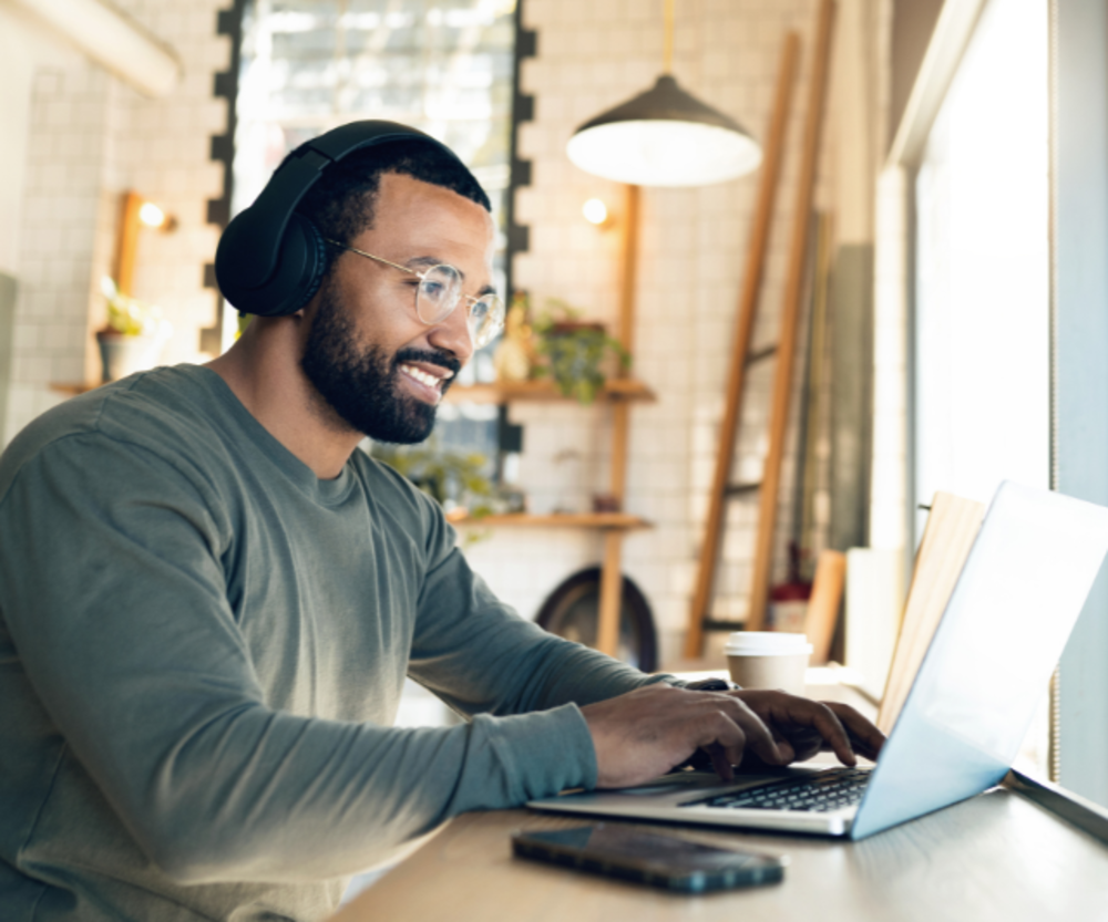Un hombre con auriculares sonriendo mientras trabaja en un ordenador, con una taza de café y un móvil junto a él.