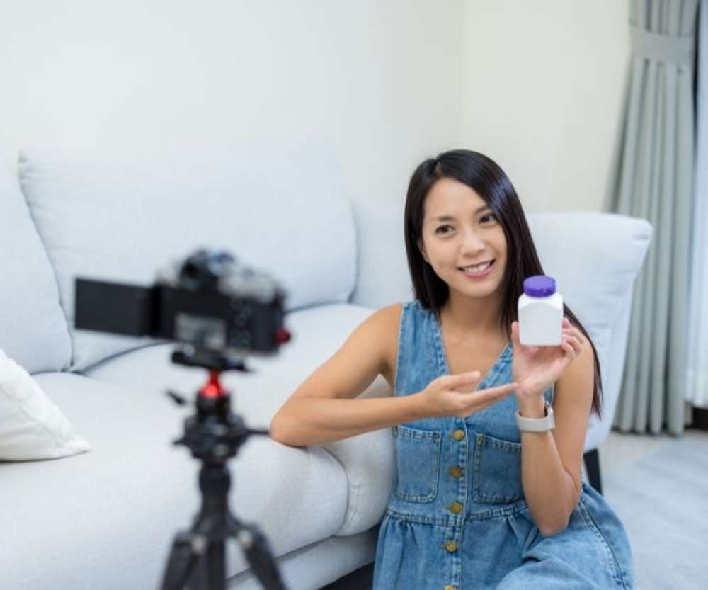 A young woman recording a video of herself holding up a supplement bottle to the camera.