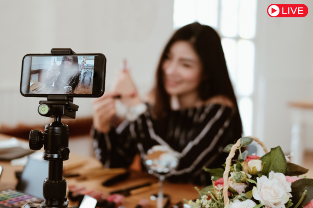 A woman live streaming while showcasing products to her audience using a smartphone and tripod setup.