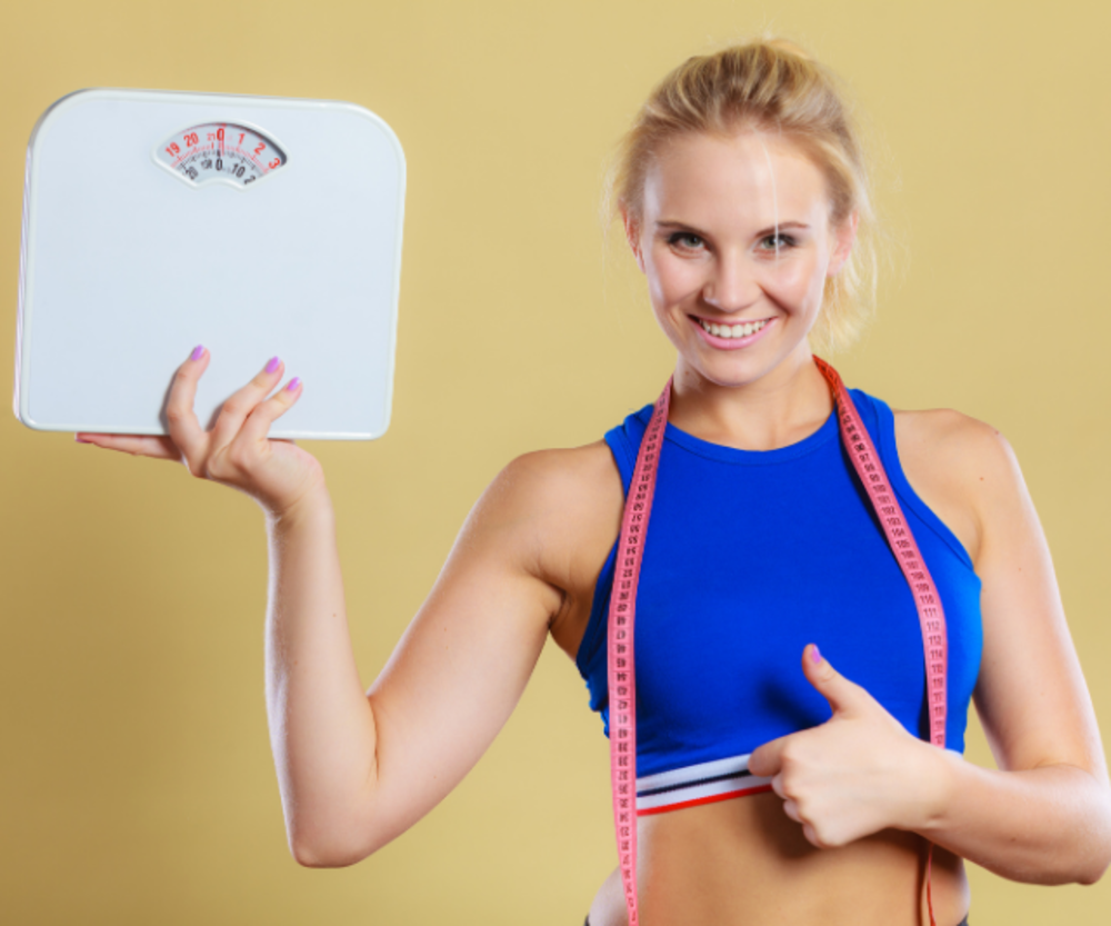 A fit woman in a blue sports bra smiling, holding a scale in one hand and a measuring tape around her neck, giving a thumbs up.