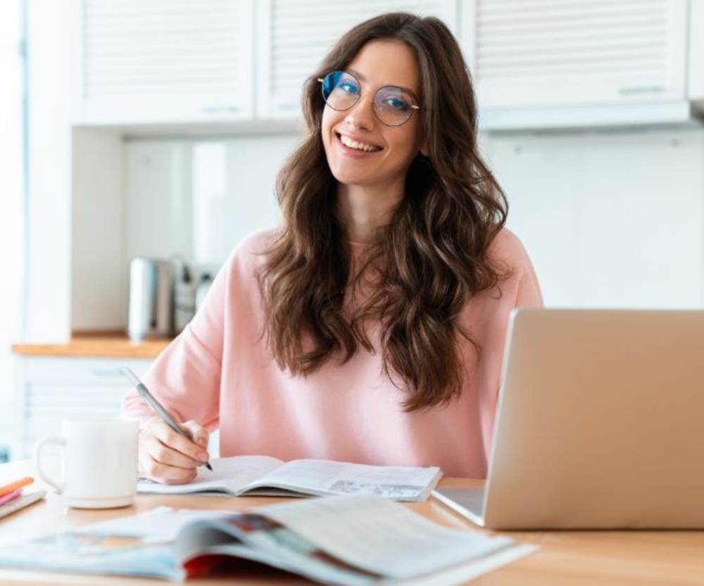 Woman in pink sweater smiling at camera, writing in a notebook at her desk with a laptop and coffee mug.