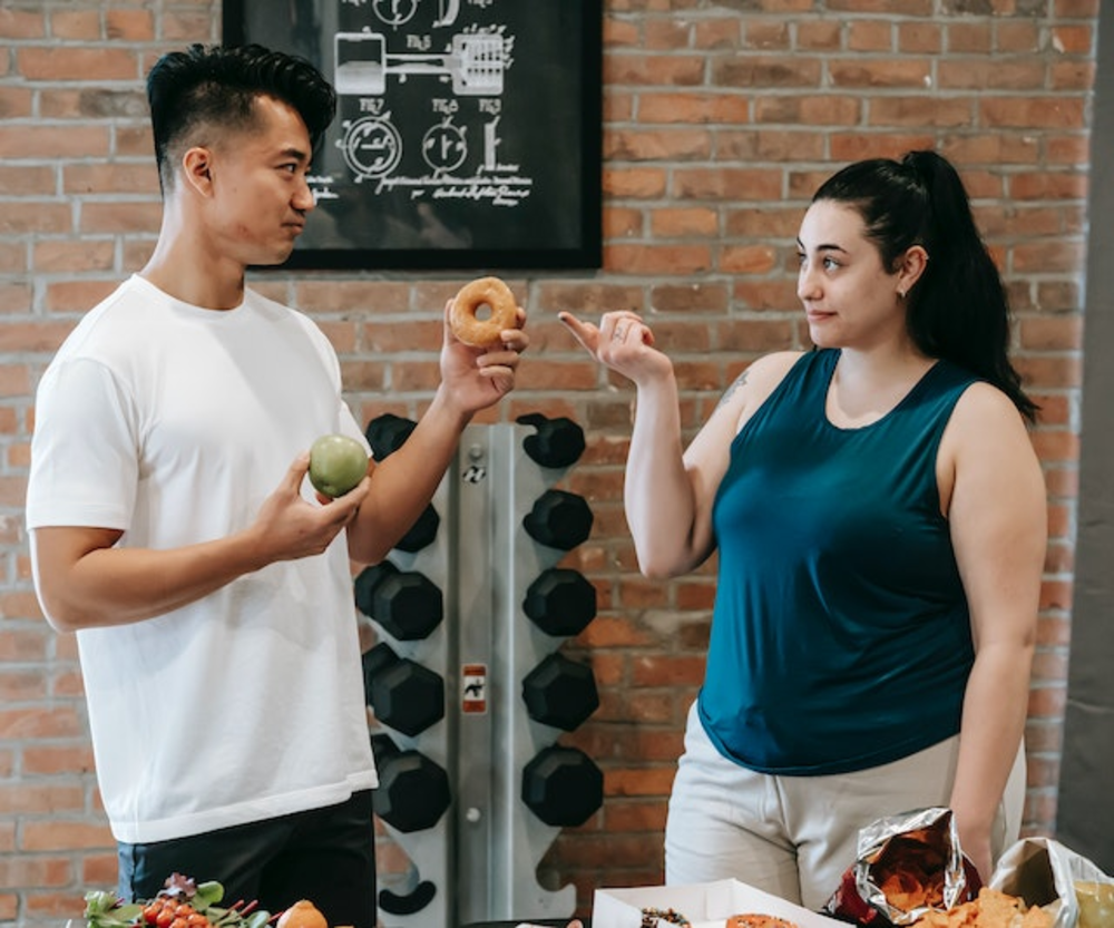 A man holding an apple and a woman pointing to a doughnut in a gym, illustrating a conversation about healthy food choices versus indulgent treats.