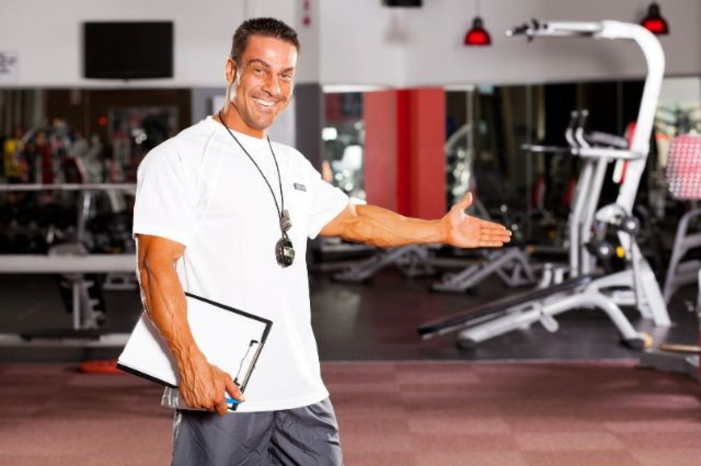 Ein lächelnder Personal Trainer im Fitnessstudio, der eine einladende Geste macht, vermutlich gegenüber einem neuen Kunden oder einer neuen Klasse.