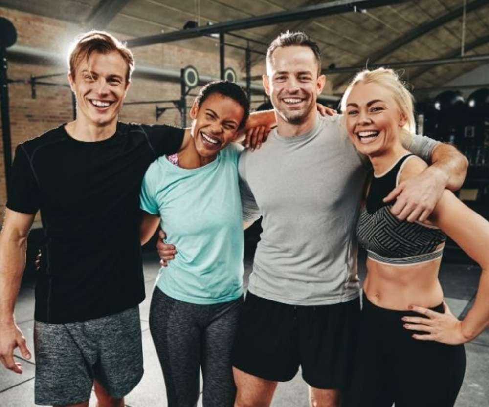 Group of four happy gym members smiling and embracing each other in a gym setting.