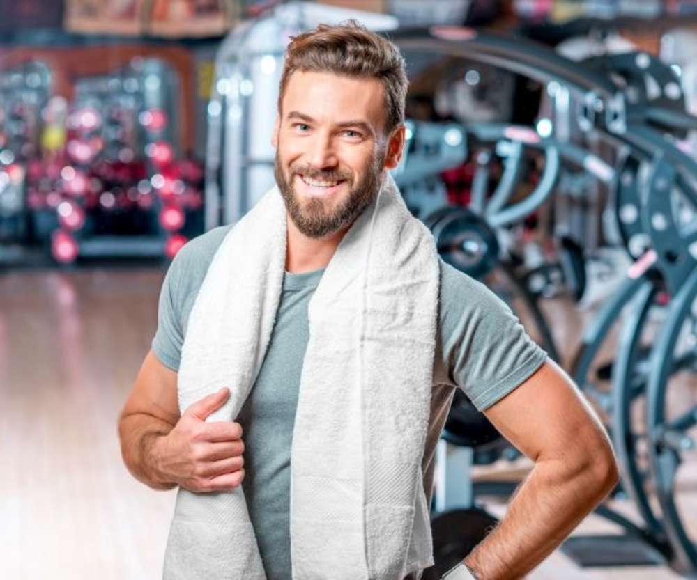 A smiling bearded man with a towel around his neck in a gym setting.