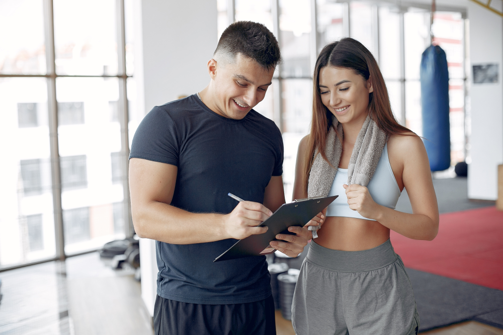 Eine Frau im Fitnessstudio, die sich mit ihrem Personal Trainer unterhält und einige Informationen durchgeht.