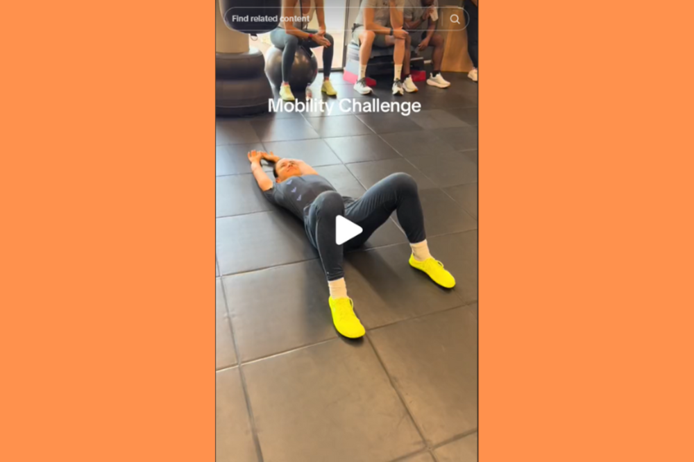 Eine Person, die an einer Mobilitätsherausforderung teilnimmt, indem sie Bodenübungen in einer Fitnessumgebung ausführt, mit Zuschauern im Hintergrund.