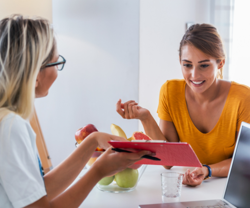 A woman in a yellow top discussing nutrition with a dietitian who is holding a tablet and a plate with fruits.