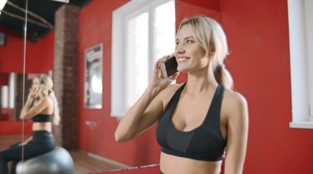 Eine Frau in Trainingskleidung telefoniert in einem Sportstudio, was darauf hindeuten könnte, dass sie ihren Zeitplan verwaltet oder nach dem Training etwas aufholen möchte.