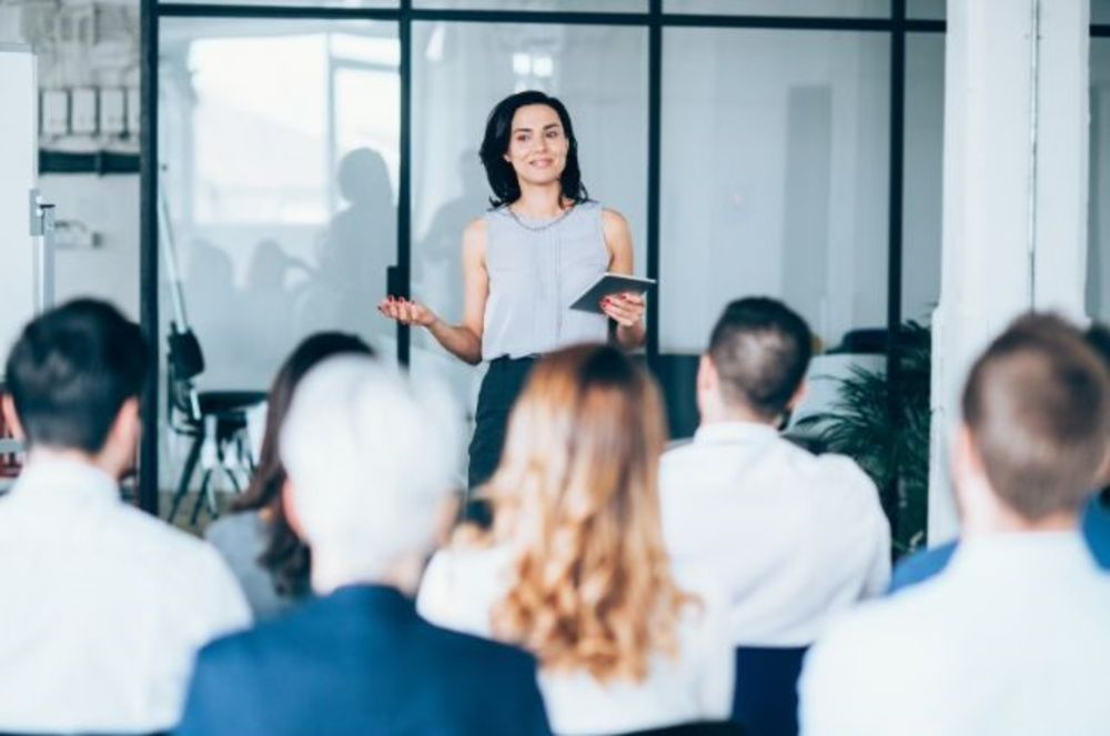 Una mujer presentando en un seminario o taller a una audiencia en un entorno empresarial.