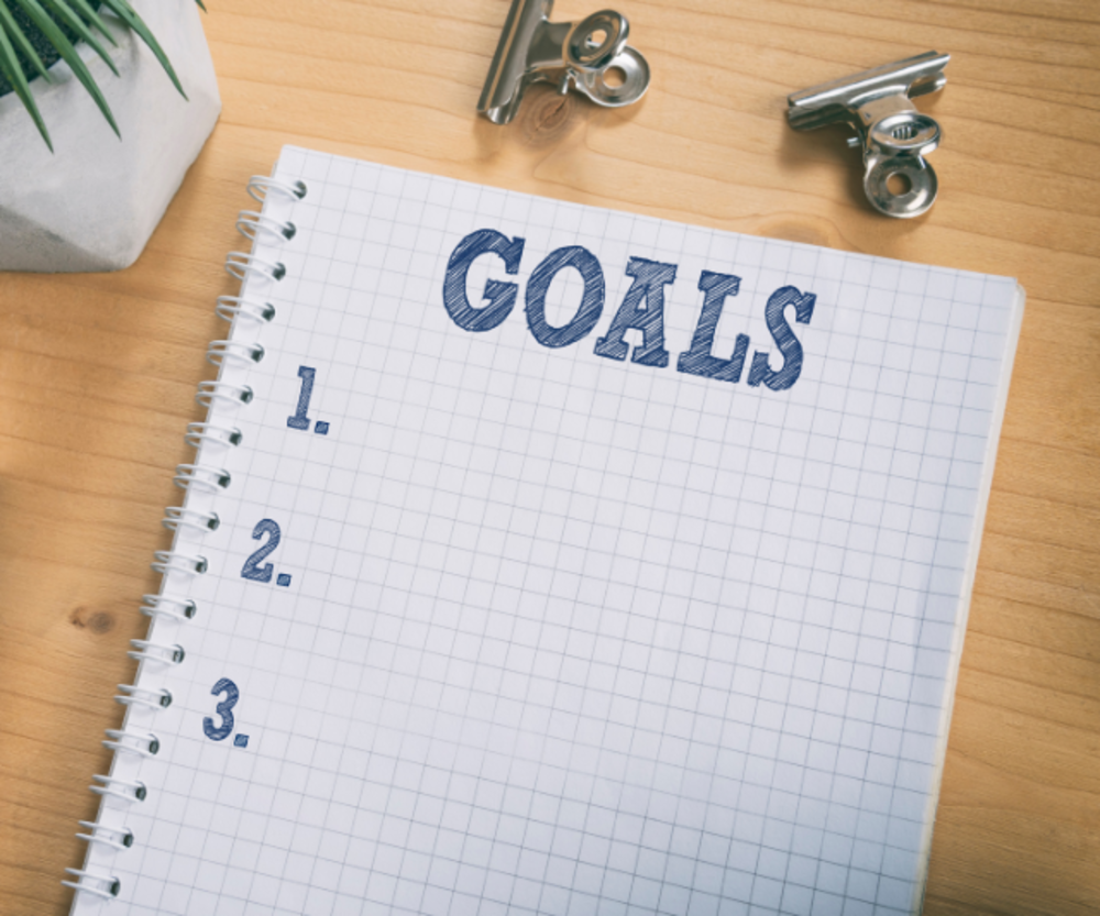 An open notebook with a grid pattern, labeled "GOALS" at the top, and a numbered list starting with 1, 2, and 3, on a wooden desk.