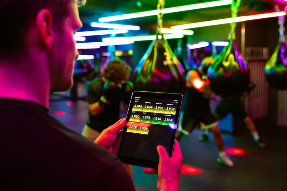 Eine Person hält ein Tablet mit Fitnessstatistiken in einem Fitnessstudio mit bunten Neonröhren.
