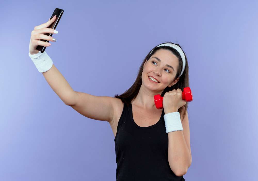 A woman in a black tank top holding up a red dumbbell and taking a selfie with a cell phone
