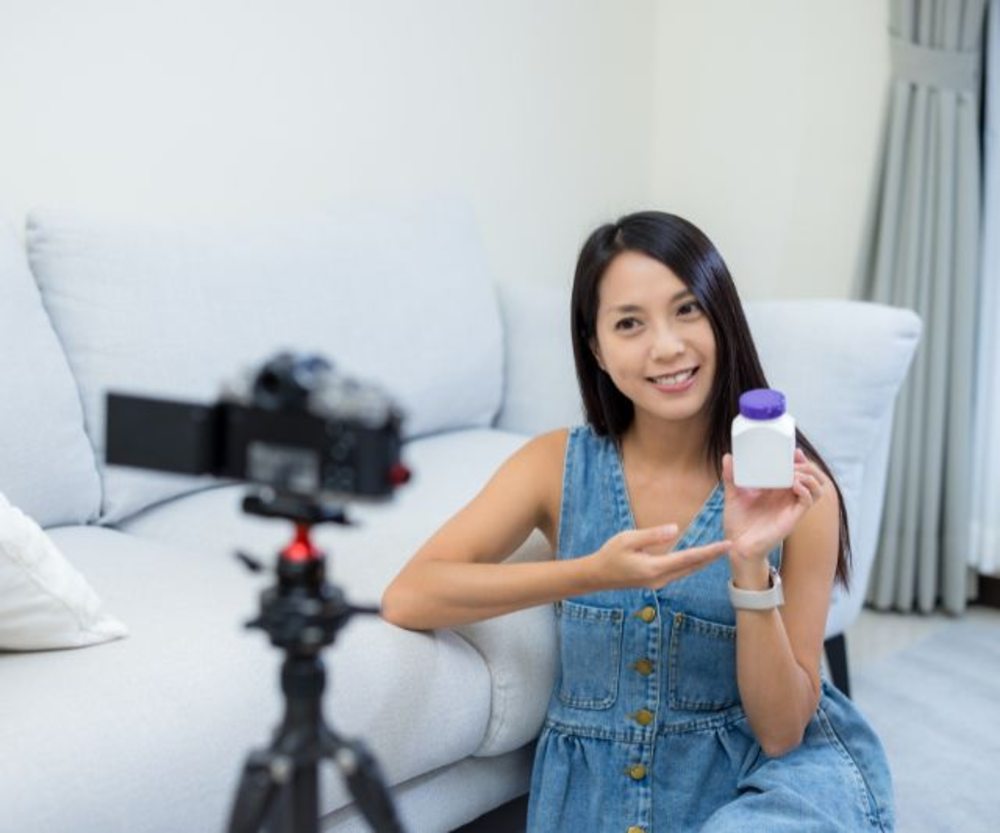 Una chica sentada en un sofá presentando una botella de suplemento, probablemente creando contenido para una reseña o recomendación de producto.