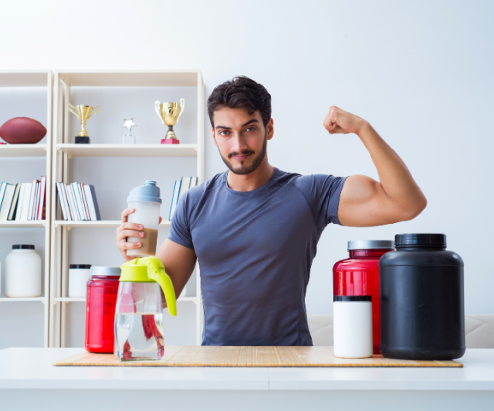 A man flexing his biceps and holding a shaker bottle, with protein supplement containers on the table in front of him.