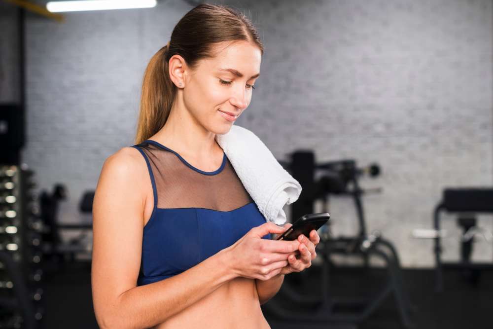 Woman in a gym checking her smartphone while holding a towel.