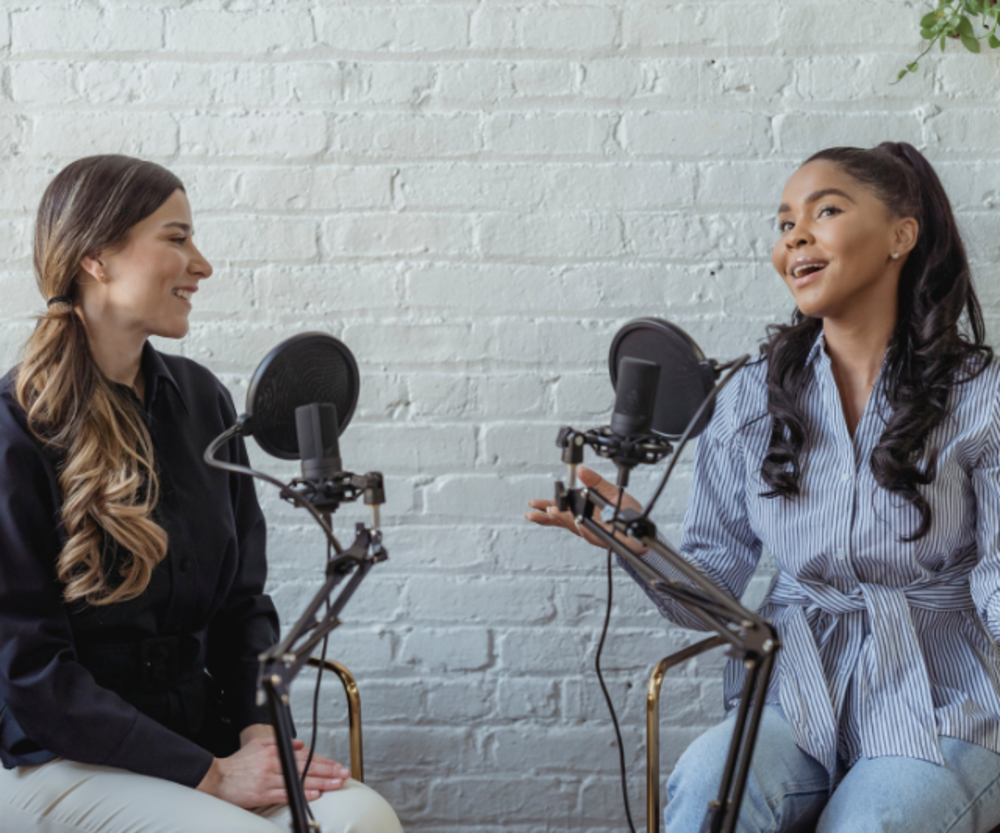 Two women engaging in a conversation while recording a podcast, seated with microphones in a studio with a white brick background.
