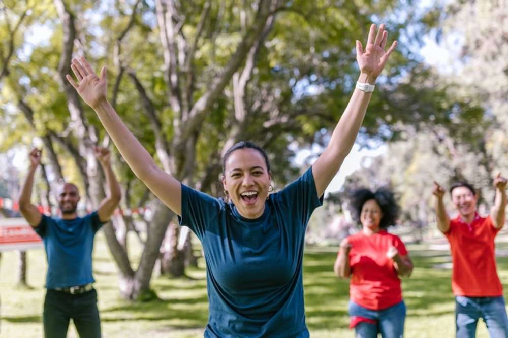 Group of happy people exercising together outdoors, with a woman in the foreground raising her arms in excitement.