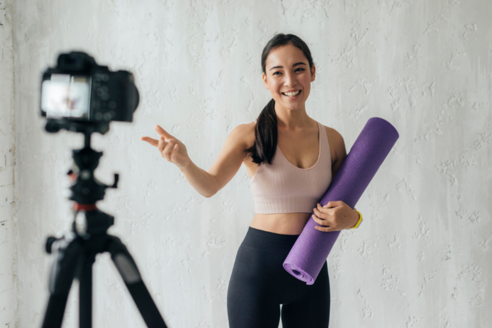 Fitness influencer smiling and holding a yoga mat while recording a video.