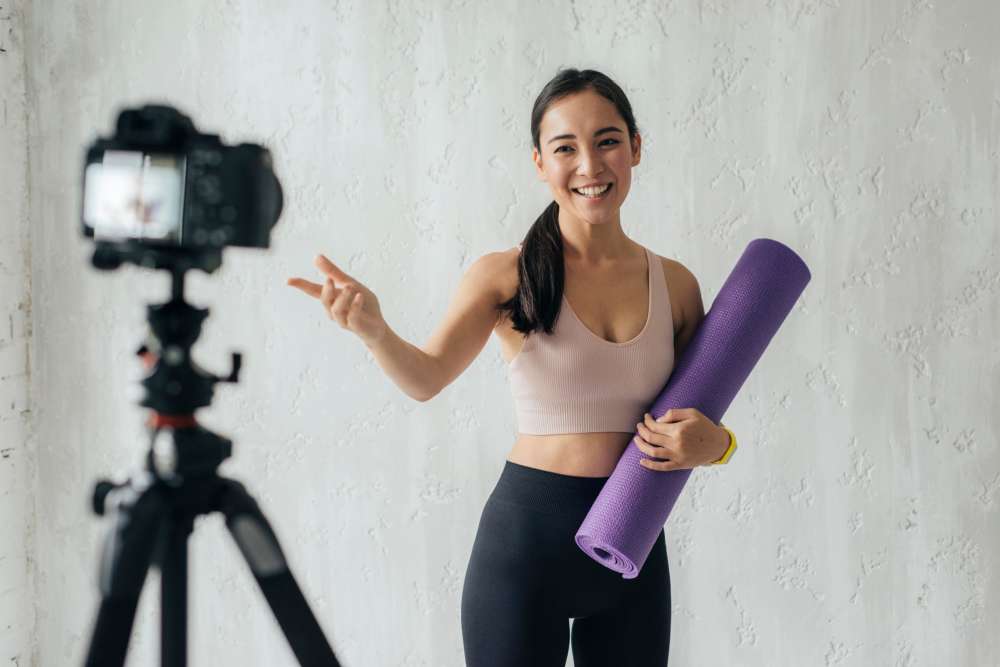 Smiling woman holding a yoga mat, speaking to a camera for a fitness video.