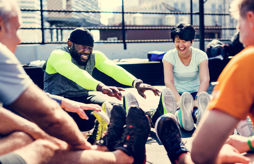 A smiling trainer in orange leads a large fitness class on a grassy field.