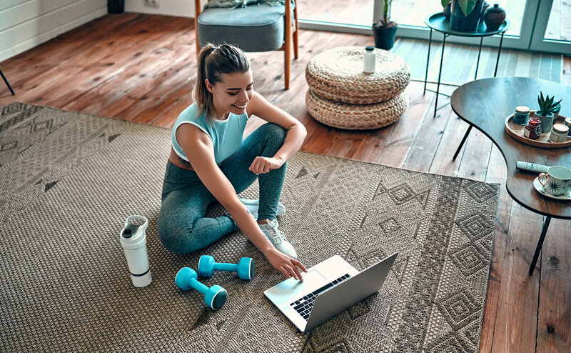 Vrouw die thuis op het tapijt zit en op zoek is naar een training op haar laptop, halters en waterfles naast haar