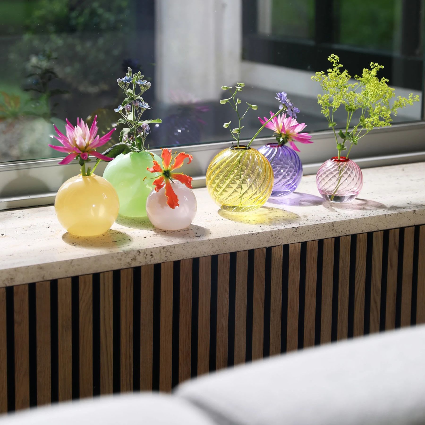 Kleurrijke Bubblegum vaasjes op vensterbank met kleine bloemen, waaronder een groene glazen bolvaas
