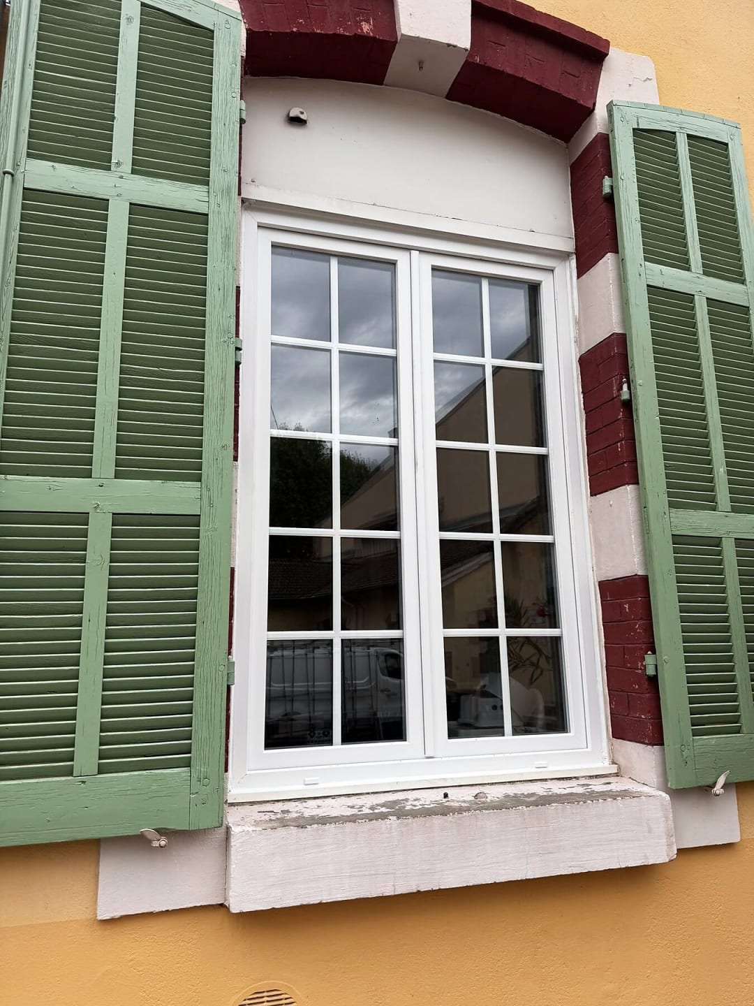 Fenêtre PVC à petits bois encadrée de volets verts sur une façade ocre