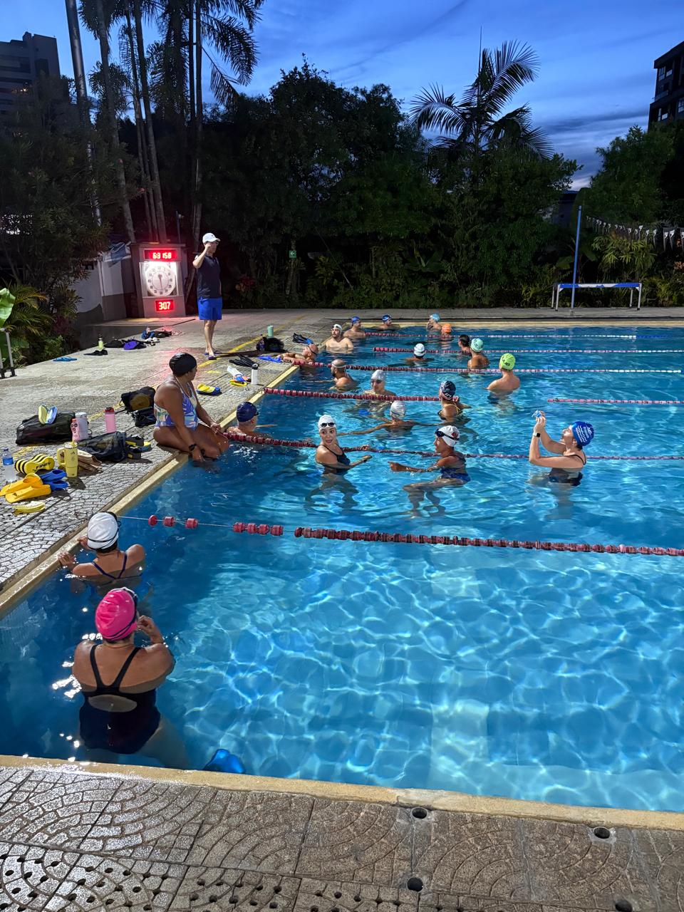 entrenador dirigiéndose a sus nadadores quiénes escuchan atentos sus instrucciones, durante una sesión de natación nocturna