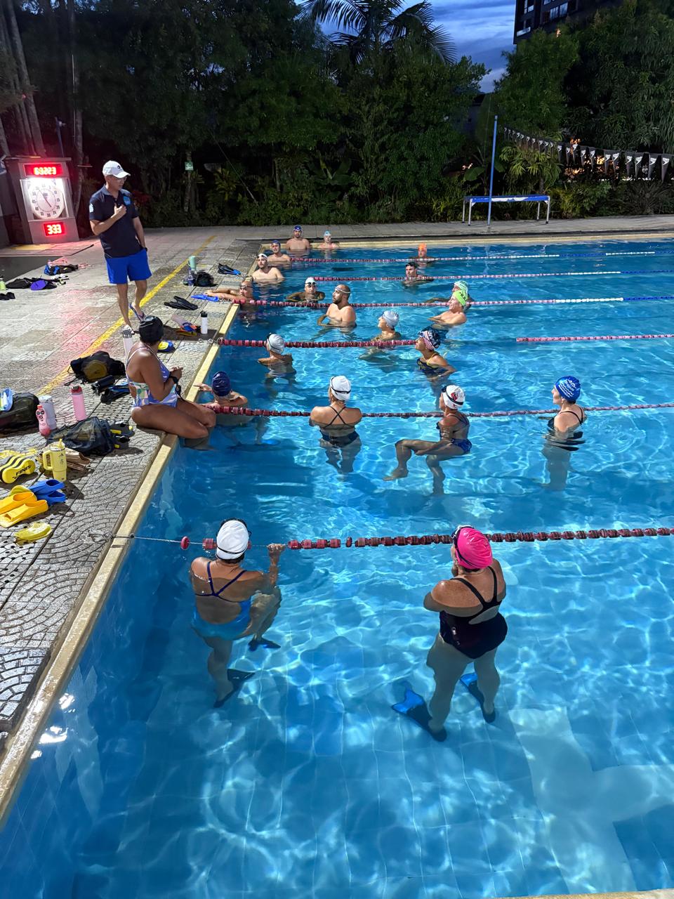 entrenador dirigiéndose a un grupo de nadadores masters, quienes escuchan atentos durante una sesión de entrenamiento nocturna